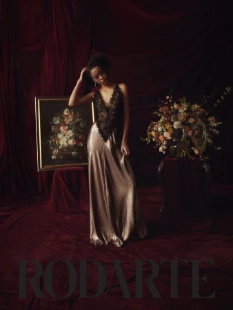 A Black female fashion model poses indoors in a gown with a lace bodice in front of a floral arrangement, vintage painting and velvet curtains.