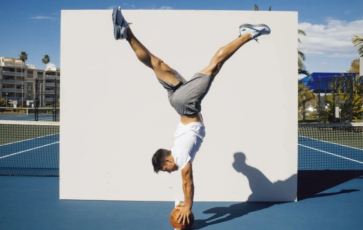 Athletic man balances in a handstand on a basketball outdoors, with feet up, on a blue court near palm trees and a white backdrop.