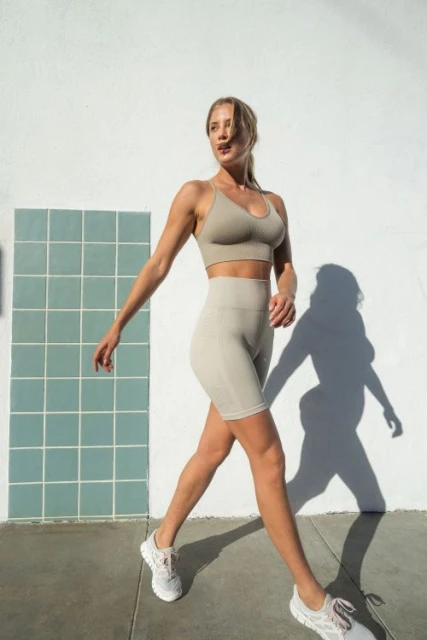 Athletic woman in exercise outfit walks near a wall. The medium shot features a sports bra, bike shorts, sneakers, and outdoor setting.