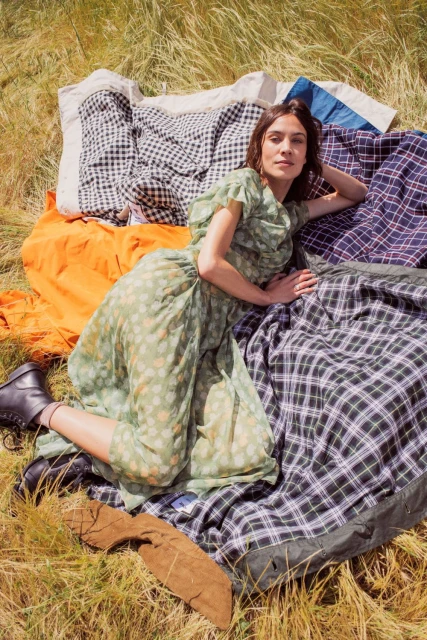 Woman in a green floral dress poses on a patchwork quilt outdoors in a field of tall grass wearing black leather boots.