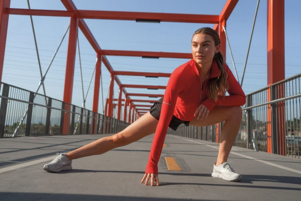 Athletic woman in red jacket performs a hamstring stretch on a pedestrian bridge, wearing running shoes, medium shot, clear sky.