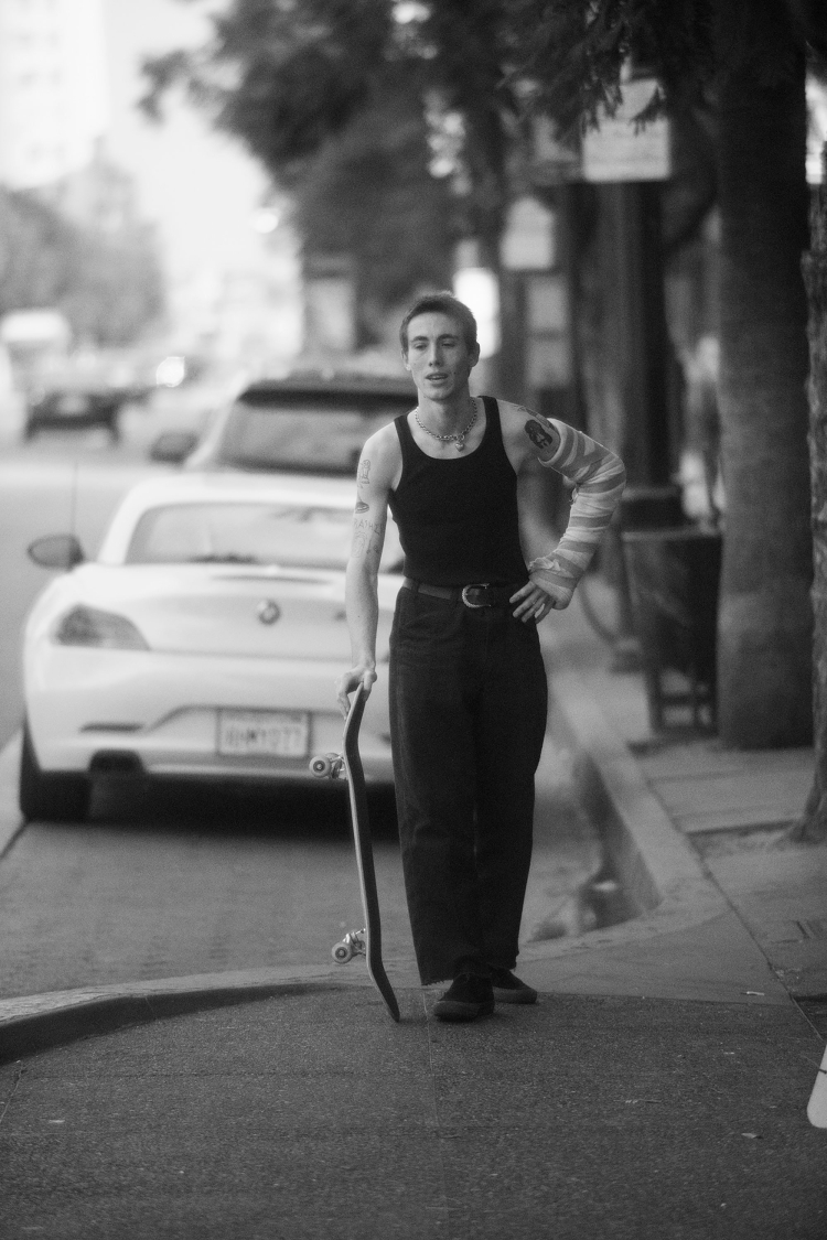 Young adult poses with skateboard outdoors, wearing a black tank top, dark pants, and an arm cast in an urban setting with a white car.