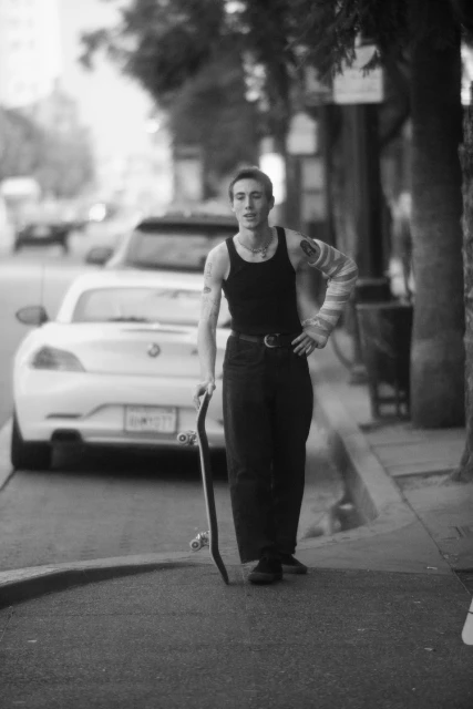 Young adult poses with skateboard outdoors, wearing a black tank top, dark pants, and an arm cast in an urban setting with a white car.