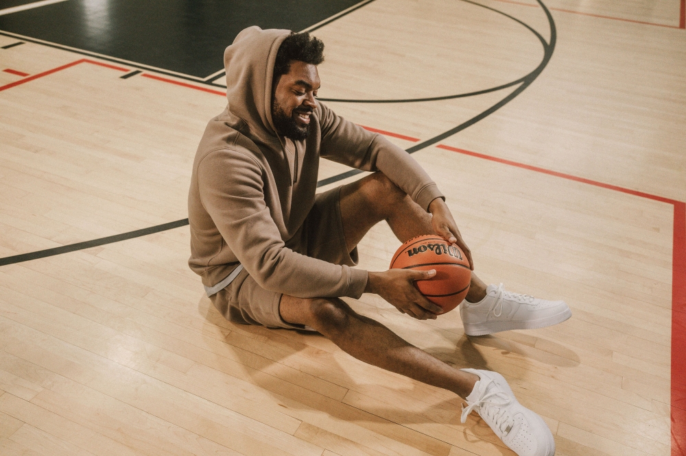 Adult male in a tan hoodie and shorts sits on a basketball court with a Wilson basketball and white sneakers in an indoor setting.