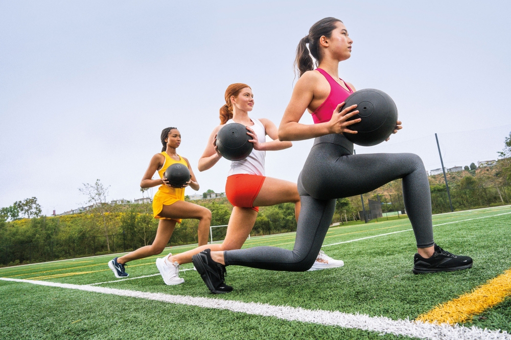 Three women in athletic wear perform lunges with medicine balls on a green field under an overcast sky, showcasing fitness and teamwork.