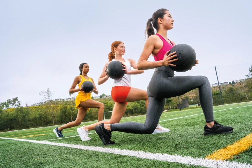 Three women in athletic wear perform lunges with medicine balls on a green field under an overcast sky, showcasing fitness and teamwork.