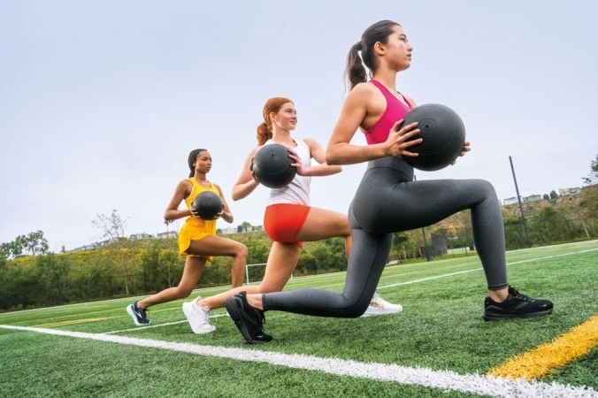 Three women in athletic wear perform lunges with medicine balls on a green field under an overcast sky, showcasing fitness and teamwork.