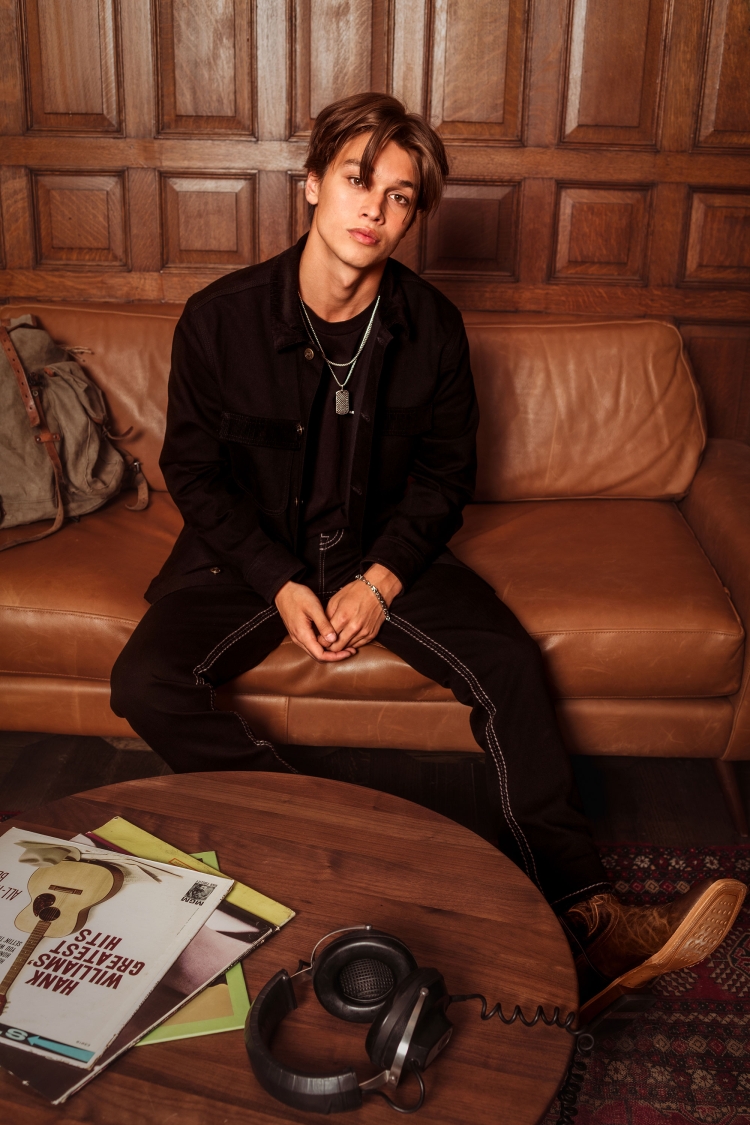 Young adult man in casual black clothing seated on a brown leather couch indoors, with headphones and records on a round wooden table.