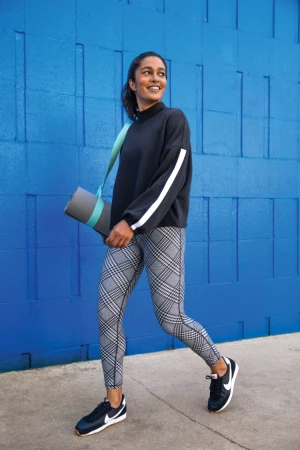 Woman walking on a sidewalk wearing a black top and patterned leggings while carrying a rolled yoga mat