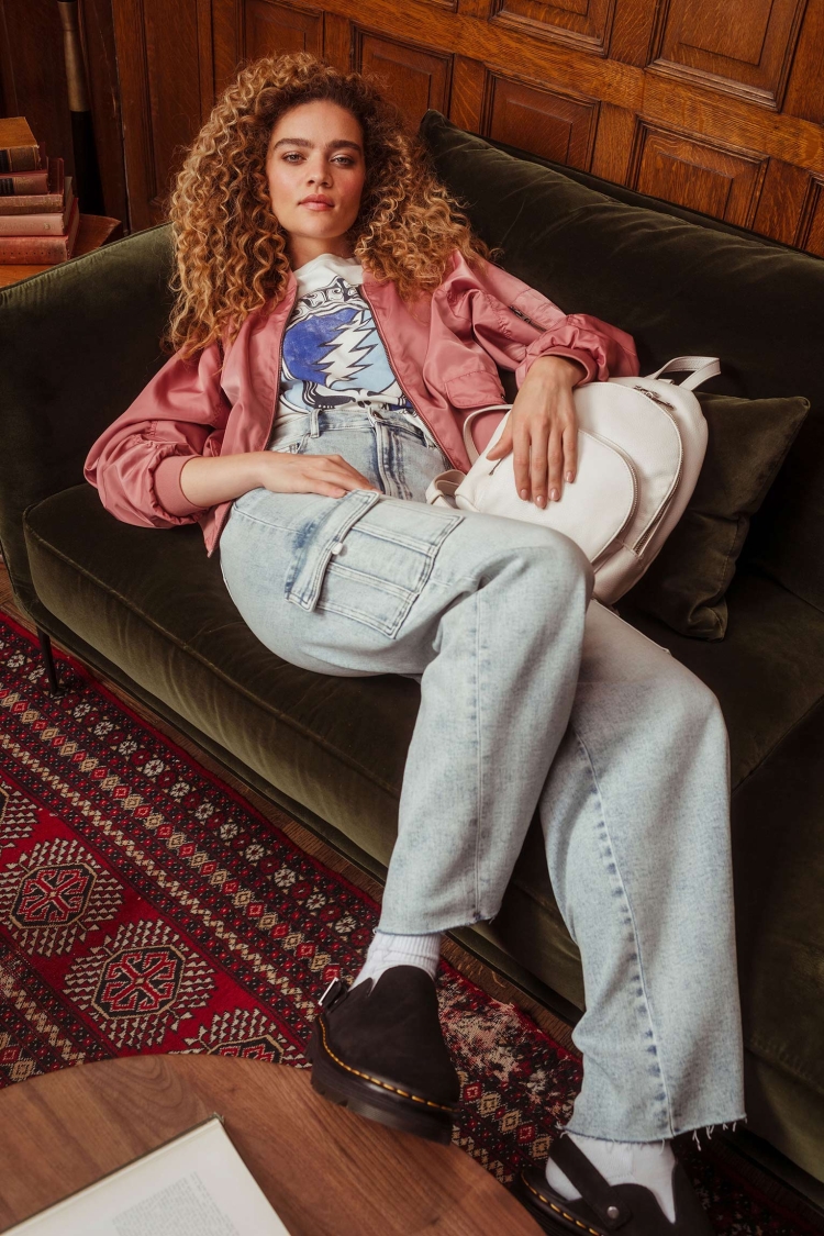 Woman with curly hair lounges on a velvet green couch, wearing a jacket, graphic tee, and jeans. Backpack and patterned rug visible.