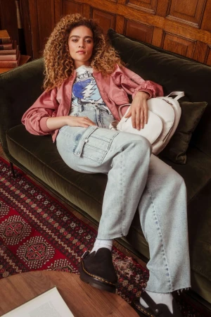 Woman with curly hair lounges on a velvet green couch, wearing a jacket, graphic tee, and jeans. Backpack and patterned rug visible.
