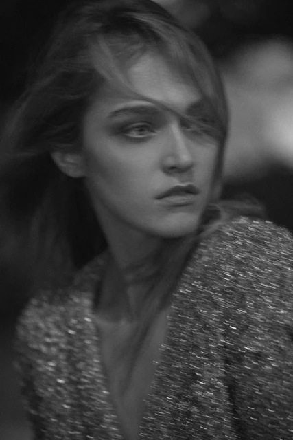 Monochrome portrait of an adult female looking to the side in a textured jacket with windblown hair against a blurry dark background.