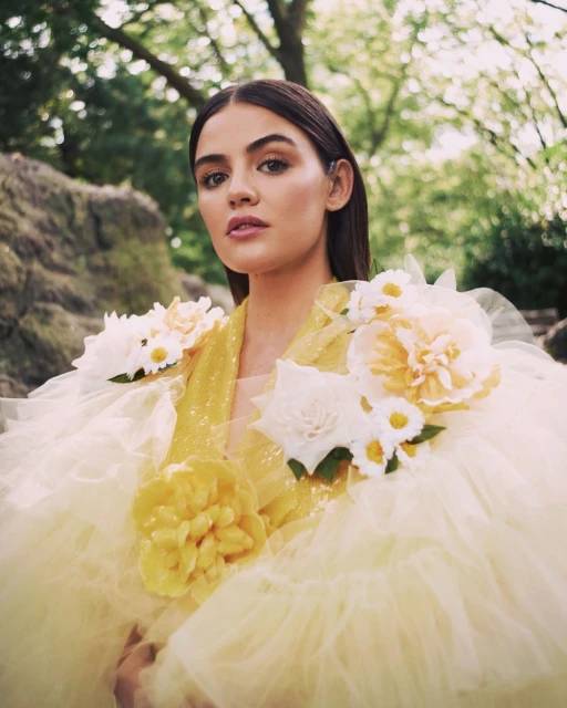 Close-up portrait of a woman in a yellow dress with flower details, set against a blurred outdoor background of greenery and rocks.