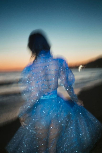 A blurry view of a person in a blue dress walking on a beach at dusk with a gradient sky and blurred motion.
