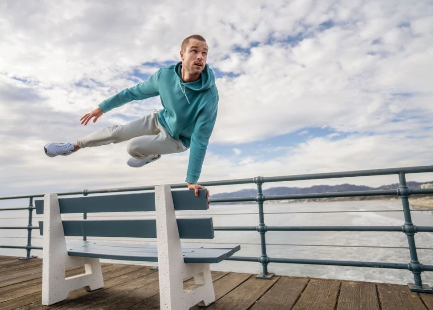 Athletic man in motion jumps on a bench, outdoors on a pier with ocean and mountains in the distance, against a cloudy sky background.