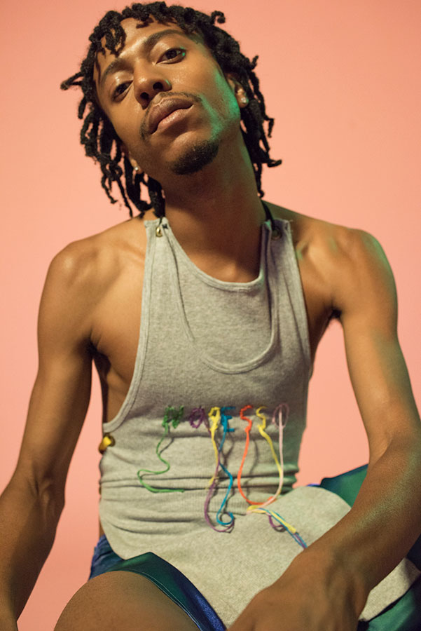 Portrait of a young adult male with dreadlocks wearing a grey tank top with embroidery against a peach-colored background in a studio.