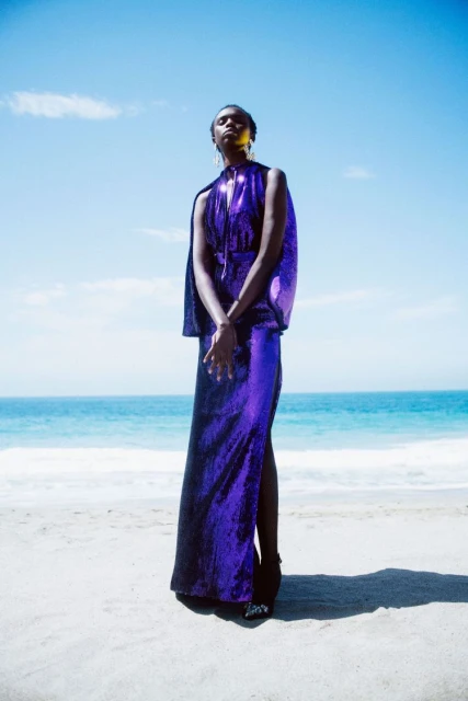 Woman in a shimmering purple dress stands on a beach with a blue ocean and sky backdrop, bathed in bright sunlight, creating a striking visual.