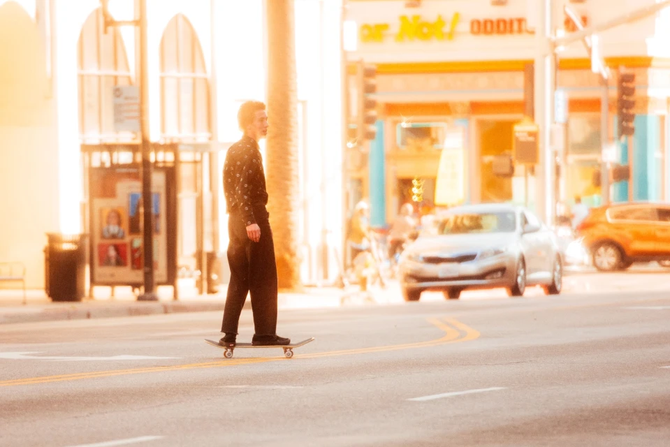 A young adult skateboarding in an urban street with cars, buildings, and bright sunlight, wearing black clothing, creating a dynamic scene.