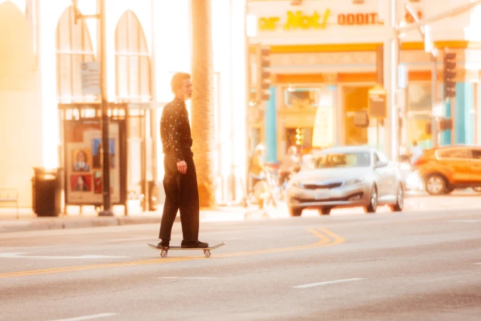 A young adult skateboarding in an urban street with cars, buildings, and bright sunlight, wearing black clothing, creating a dynamic scene.