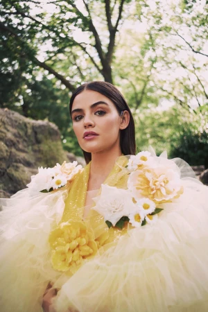 A woman wearing a yellow tulle dress adorned with large white and yellow flowers, posed outdoors beneath trees.