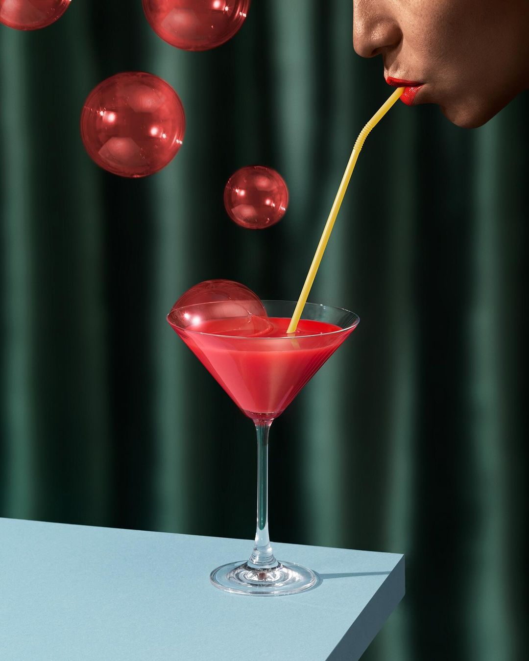 Close-up of a person drinking a red cocktail with a yellow straw against a dark green background with red transparent balls.