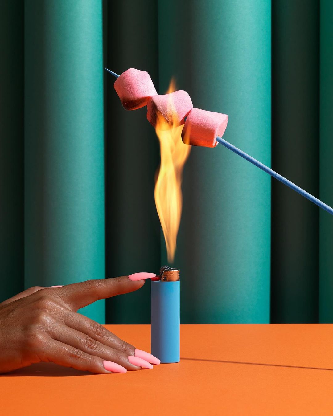 A hand holding a blue lighter under a stick of marshmallows on fire against a teal and orange background in a studio.