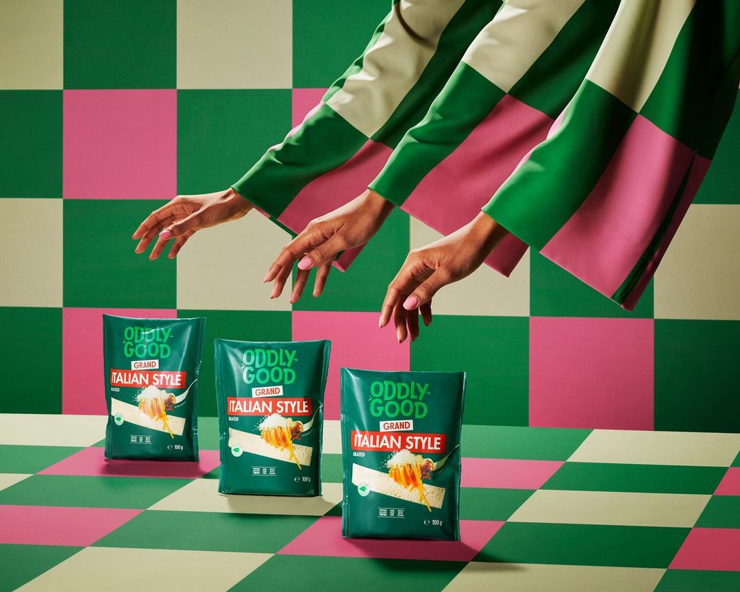 Three packages of Oddlygood Italian Style are displayed with hands reaching from above, against a green, pink, and tan checkered backdrop.