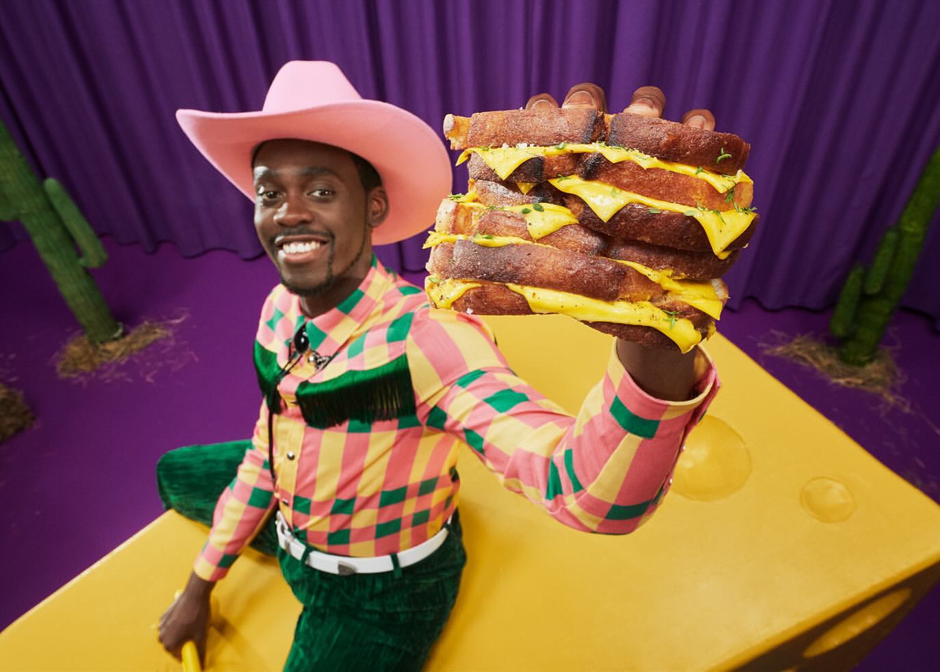 Smiling man in cowboy hat holds a stack of grilled cheese sandwiches in a vibrant studio setting with a purple backdrop.
