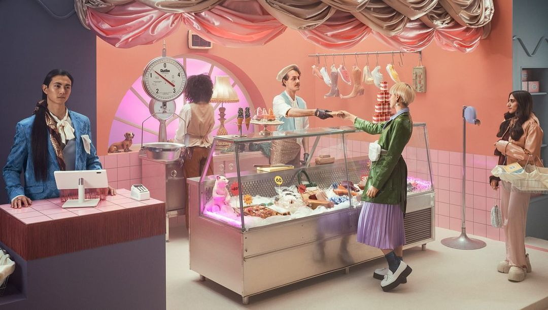Retro-styled scene with five people inside a pink-and-blue shop, featuring unique 'food' items under glass, pink tile, and whimsical decor.