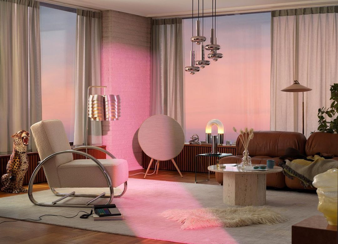 Living room interior with a beige armchair, round speaker, and modern lighting fixtures against a backdrop of a sunset-colored sky.