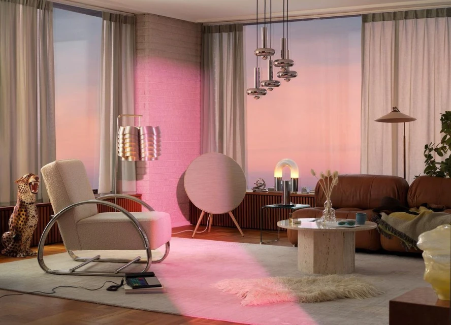Living room interior with a beige armchair, round speaker, and modern lighting fixtures against a backdrop of a sunset-colored sky.