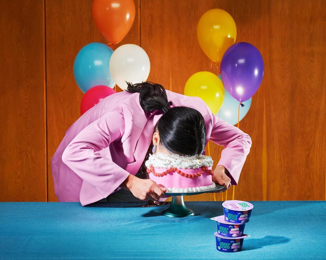 Person face-planting into pink cake with balloons in background and yogurt cups on table, creating a funny, celebratory scene.