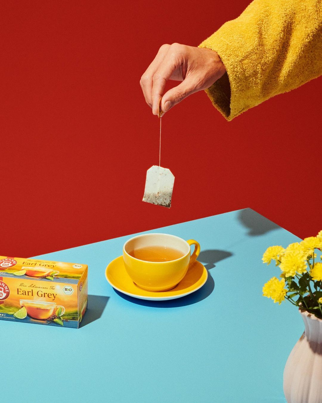 Hand holding a tea bag above a yellow cup of tea with a box of Earl Grey tea and yellow flowers, set against red and blue background.