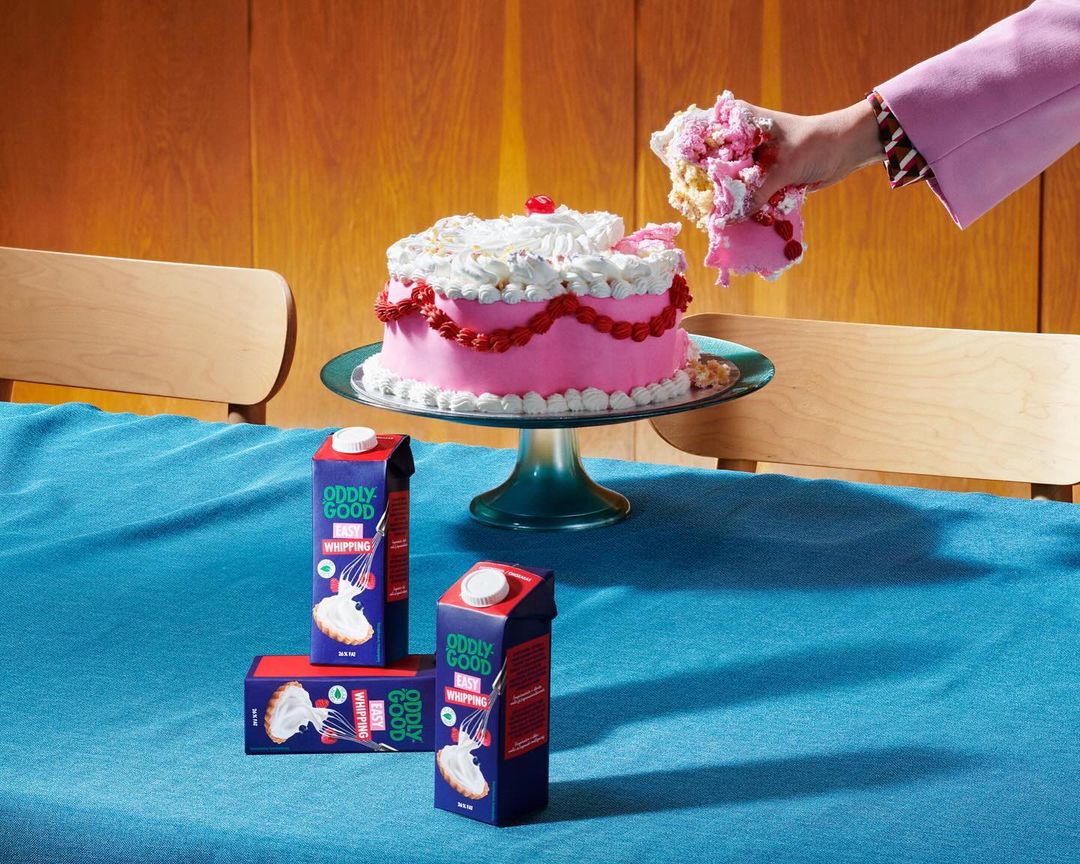 Pink frosted cake on a green cake stand with three cartons of whipping cream, one held by a person with a pink sleeve.