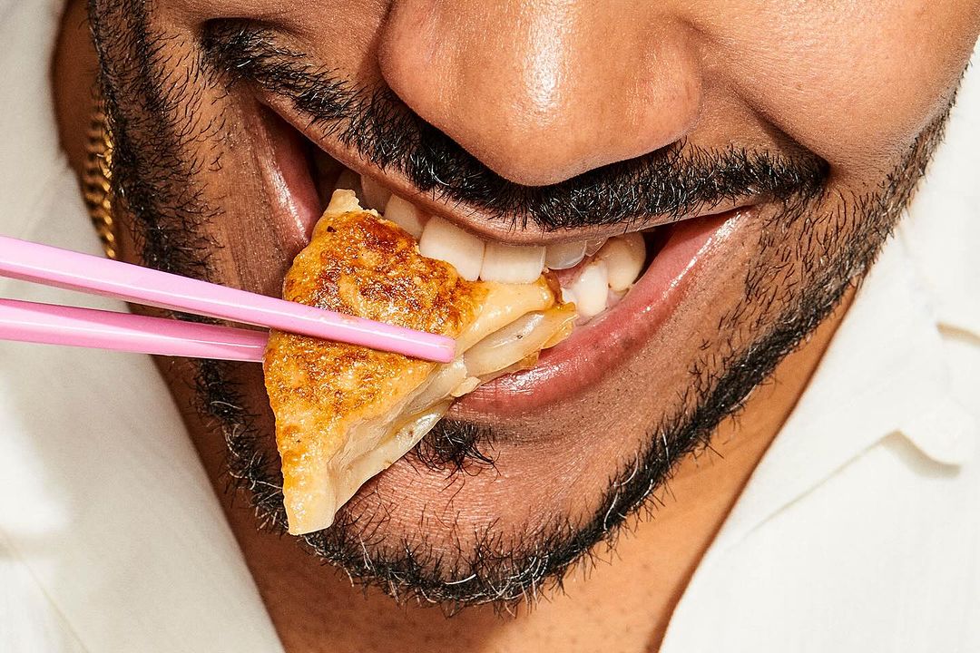 Close-up shot of a bearded adult eating pan-fried dumpling with pink chopsticks, focusing on the food and the person's mouth area.