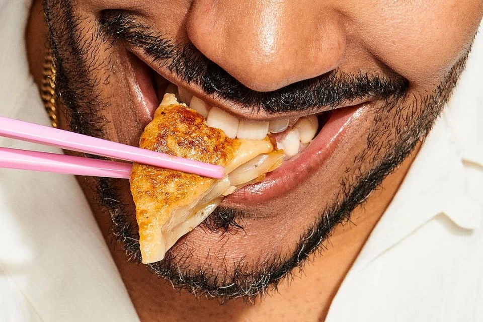 Close-up shot of a bearded adult eating pan-fried dumpling with pink chopsticks, focusing on the food and the person's mouth area.