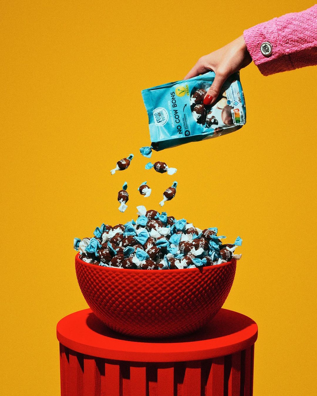 Hand pours vegan chocolate bonbons from a blue package into a red bowl on a matching red pedestal against a vibrant yellow backdrop.