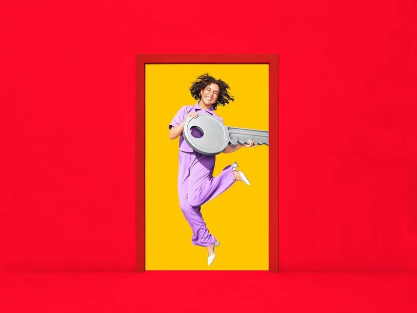 A happy woman in lavender loungewear jumps through a red doorframe holding an oversized key, set against a bright yellow background.