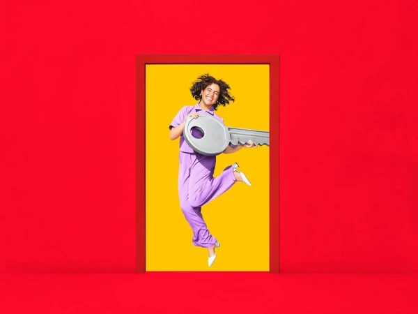 A happy woman in lavender loungewear jumps through a red doorframe holding an oversized key, set against a bright yellow background.