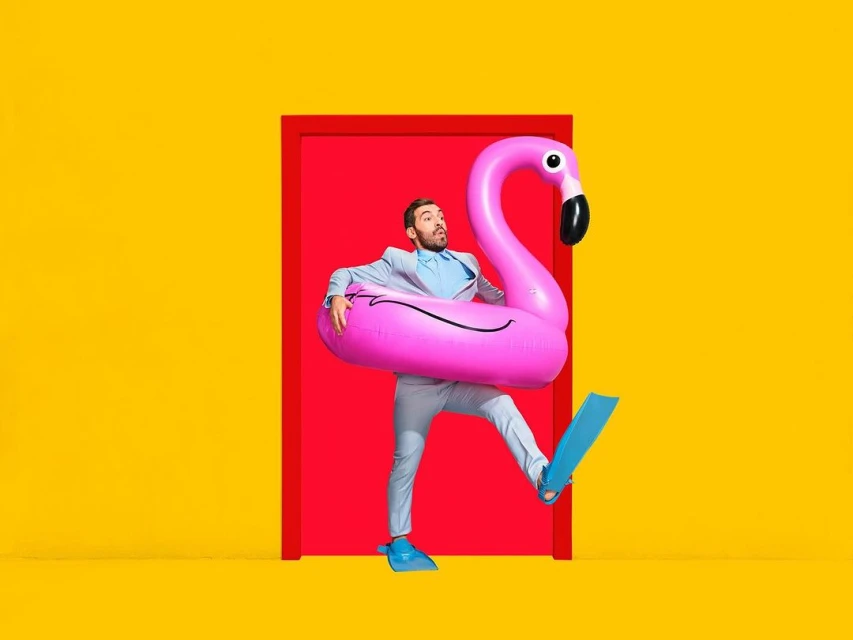 Man in suit exits red door holding pink flamingo inflatable, wearing blue flippers, against a bright yellow background in studio shot.