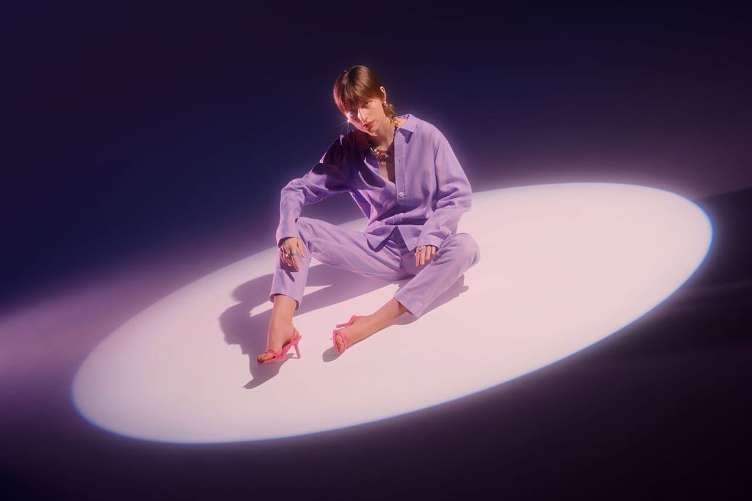 Woman in a lavender pantsuit sits in a spotlight, looking at the camera. The image evokes a fashion or beauty editorial.