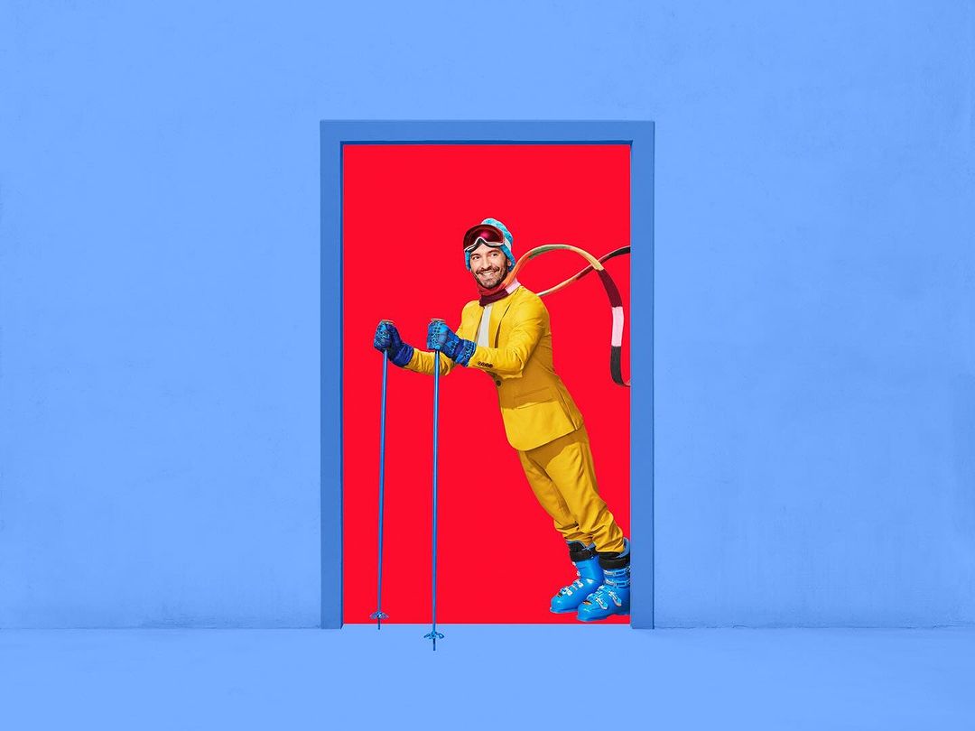 Smiling man in yellow ski suit and goggles stands in a blue doorway with a red background, holding ski poles, and wearing blue ski boots.