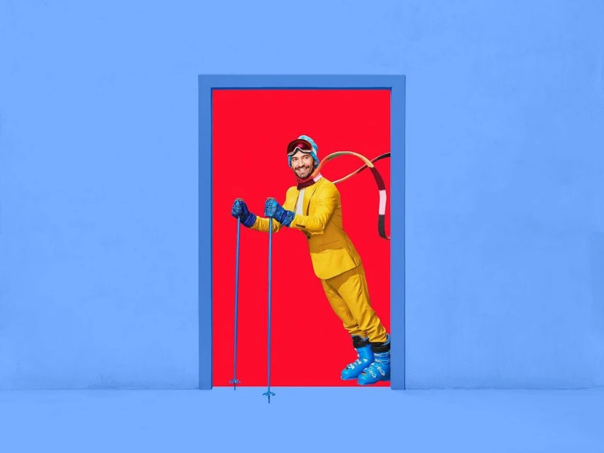 Smiling man in yellow ski suit and goggles stands in a blue doorway with a red background, holding ski poles, and wearing blue ski boots.