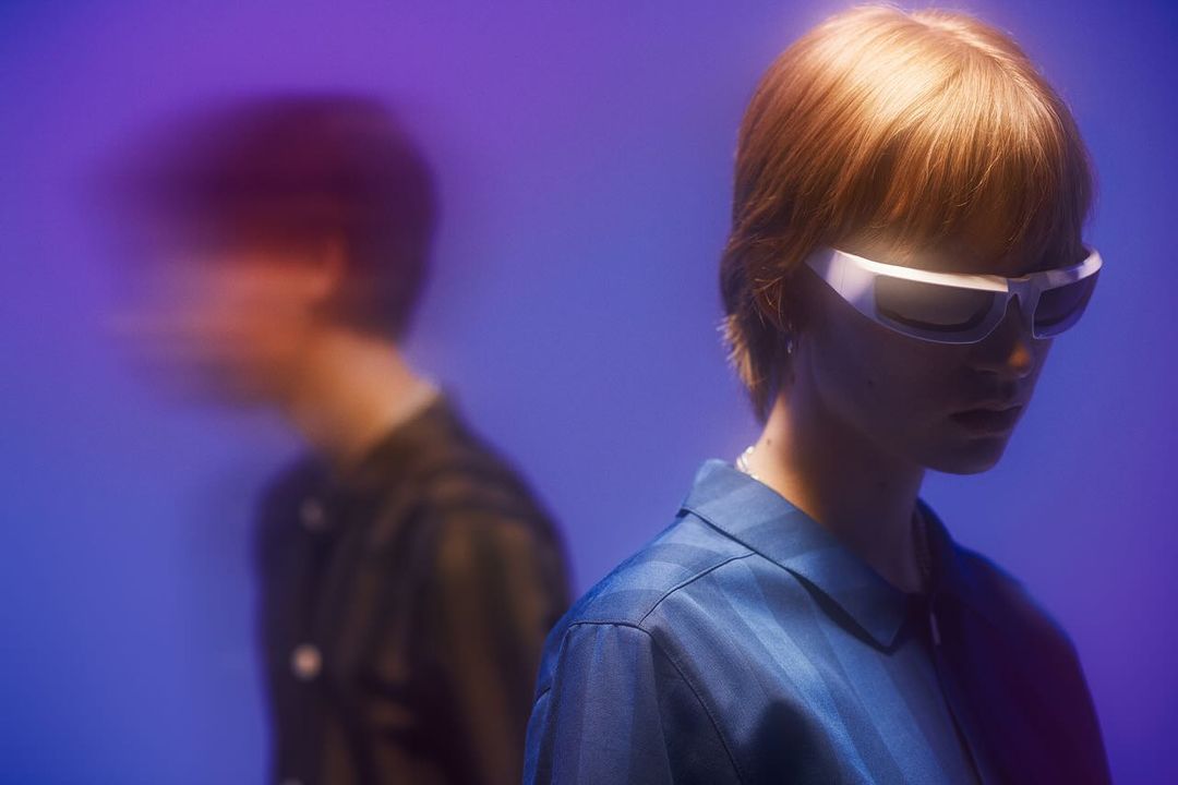Two people in a photo studio, one person in focus with futuristic sunglasses and a blue striped shirt, the other blurred in the background.