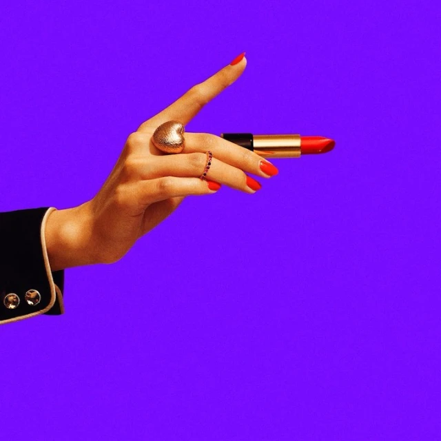 Close-up of a hand in a black blazer holding red lipstick against a vibrant purple background, also featuring rings on the fingers.