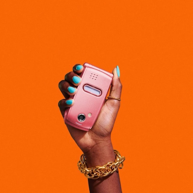 Close-up of a dark-skinned hand holding a pink flip phone with turquoise nails, a gold ring and a gold chain bracelet against an orange background.
