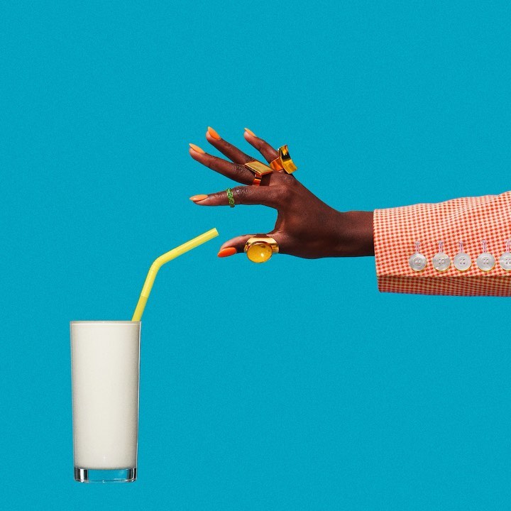 Dark-skinned hand with rings and orange nails holding a yellow straw above a glass of white liquid against a blue background.