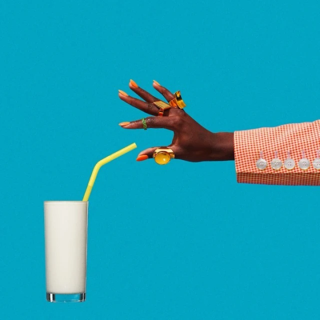 Dark-skinned hand with rings and orange nails holding a yellow straw above a glass of white liquid against a blue background.