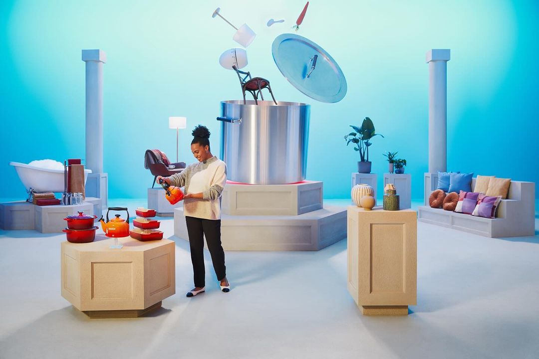 Woman adds to a colorful cookware set, with a large pot and flying kitchen utensils, in a modern setting with blue background.