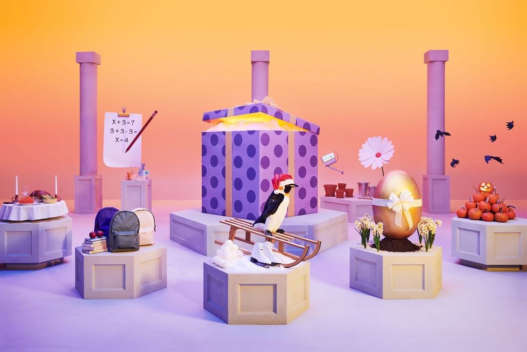 Whimsical still life featuring a toy penguin on a sled, backpacks, an Easter egg, and a school paper, against a gradient orange backdrop.